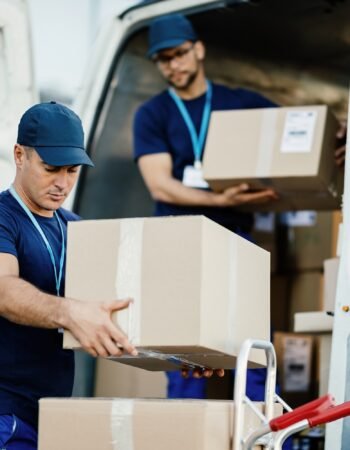 Young courier and his colleague unloading cardboard boxes from delivery van.